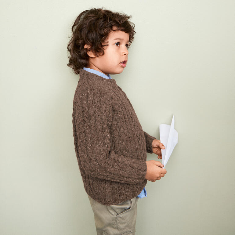 Junge in braunem Kinderpullover aus Tweed Recycled hält ein Blatt Papier.