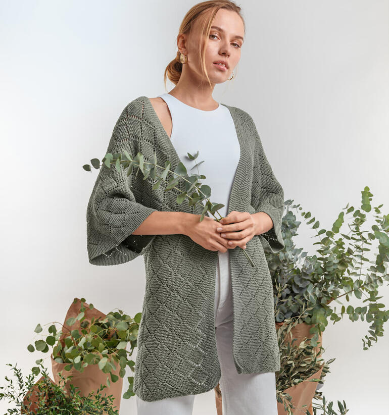 Frau trägt eine olivgrüne Kimono-Jacke aus Eco Vita, umgeben von Pflanzen.