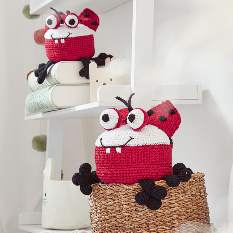 Zwei gehäkelte Marienkäferfiguren in einem Regal, eine sitzt auf einem Korb.