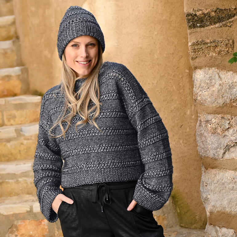 Frau trägt grauen Kurzpullover und Mütze, steht an einer Steinwand.