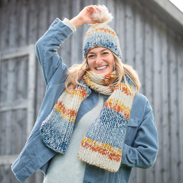 Lächelnde Frau trägt Schal und Mütze aus Nordic Chunky in bunten Farben vor einer Holzscheune.