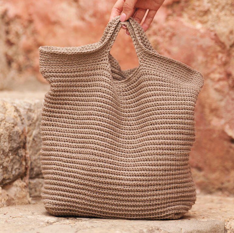 Braune Häkeltasche aus Creative Linen wird gehalten mit einer steinernen Wand im Hintergrund.