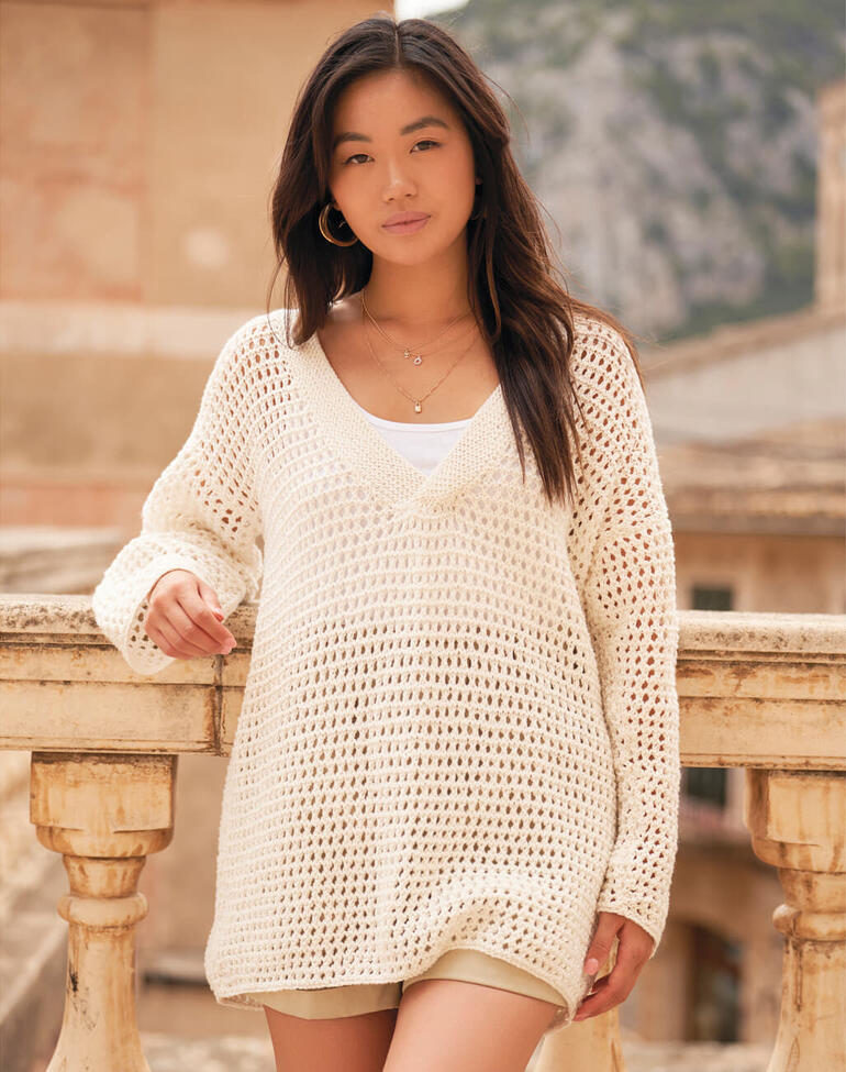 Frau trägt einen grobmaschigen Pullover in cremefarben, posiert auf einem Balkon mit Blick auf eine alte Stadt.