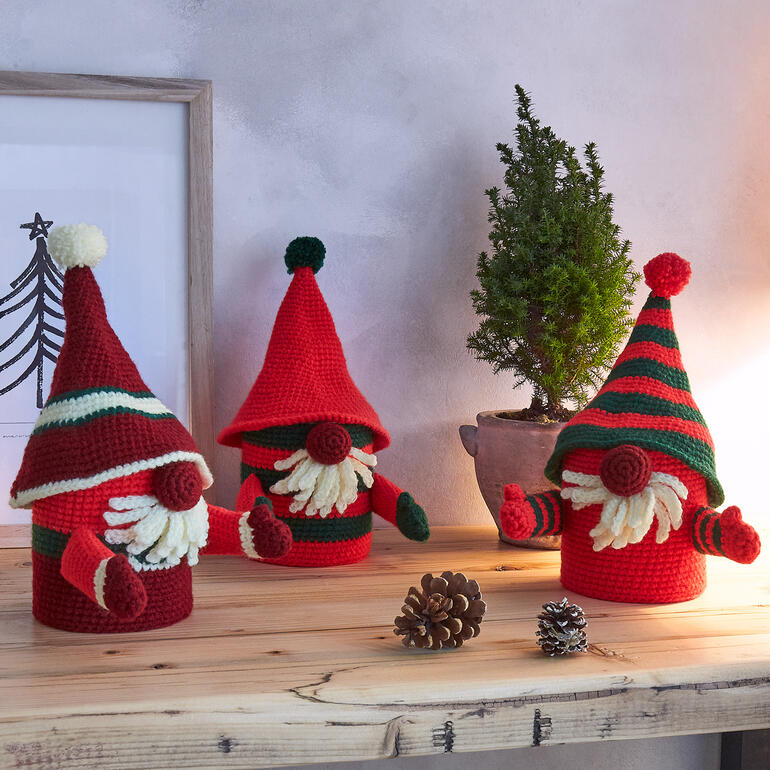 Drei Wichtel-Weihnachtsdosen, gehäkelt in Rot und Grün, stehen auf einem Tisch dekoriert mit Tannenzapfen.
