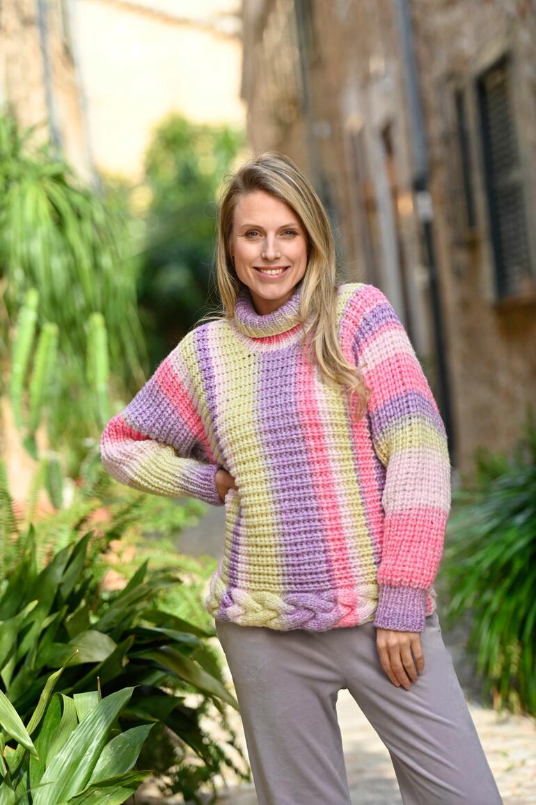 Frau mit buntem, quergestricktem Pullover posaunt voller Freude in einem Garten.