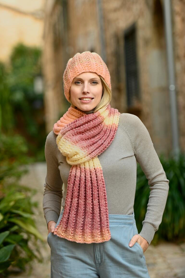 Frau trägt einen Schal und eine Mütze aus Linie 519 Cosmo, posiert lächelnd auf einem gepflasterten Weg.