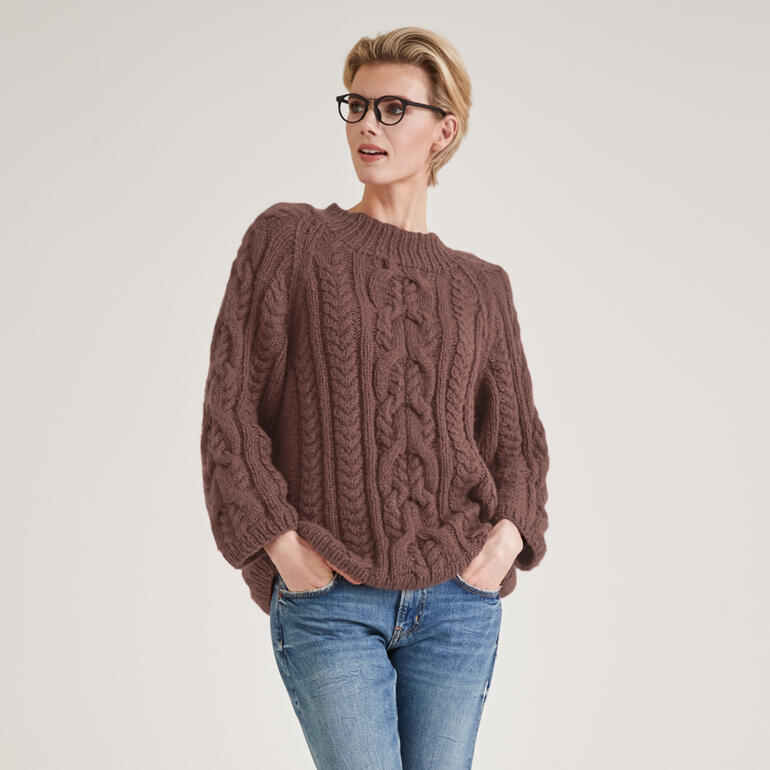 Frau trägt einen braunen Pullover aus NewWool, posiert mit Brille in neutralem Licht.