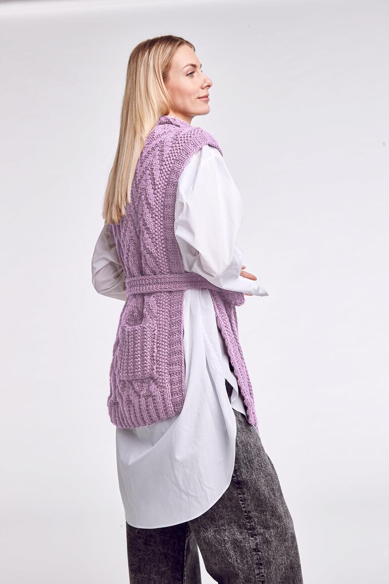 Frau in langer lila Strickweste posiert von der Seite in einem Studio.