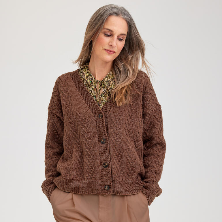 Frau trägt eine braune Jacke aus Aparta-Nova, lächelt und posiert in einem hellen Raum.