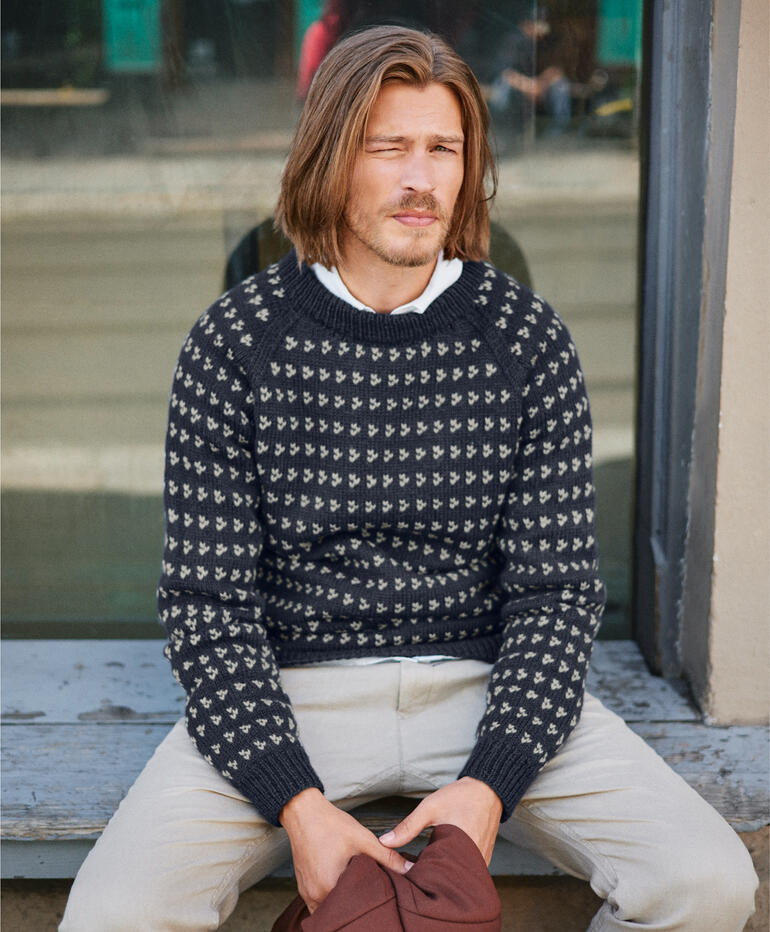 Mann sitzt draußen in einem gestrickten, gemusterten Pullover.