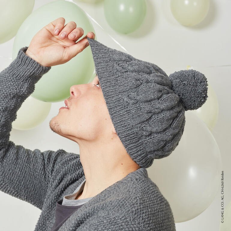 Mann zieht eine graue Lambswool-Mütze mit einem Bommel auf, steht vor Luftballons.