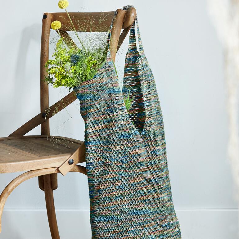 Häkeltasche aus buntem Garn hängt an einem Stuhl, mit Blumen gefüllt.