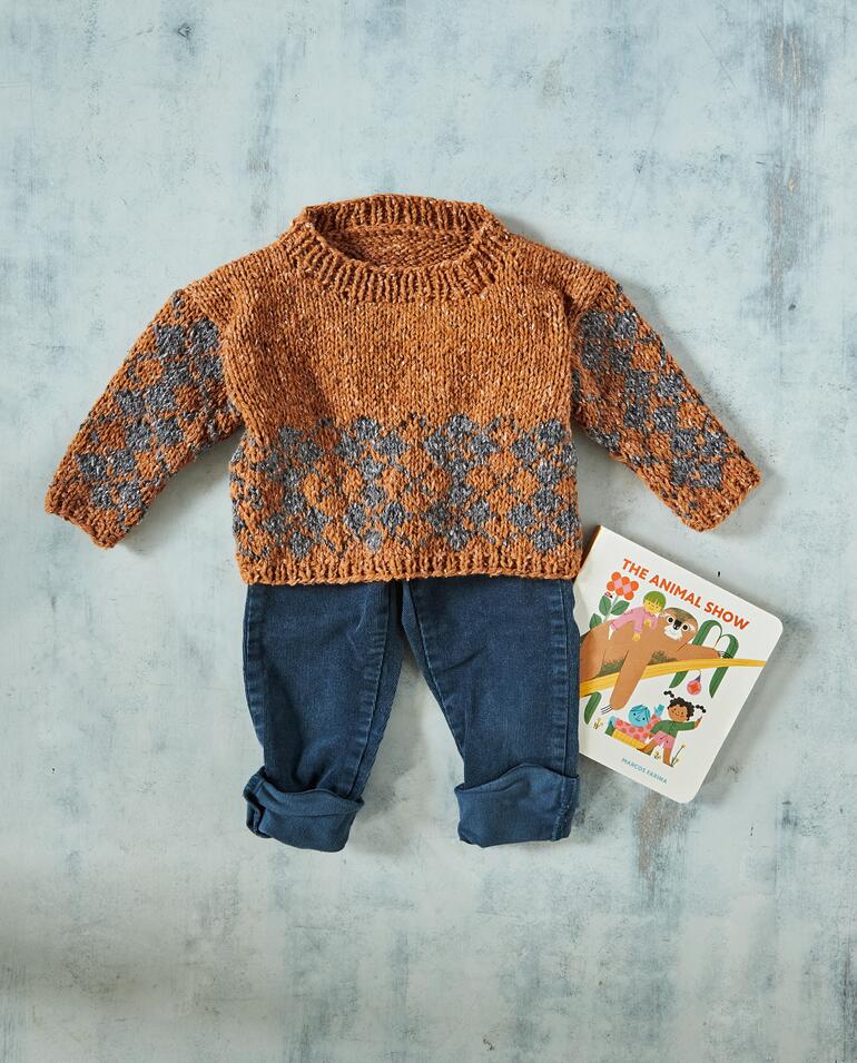 Häkelpullover für Babys in warmen Farben, liegt über kleinen Jeans.