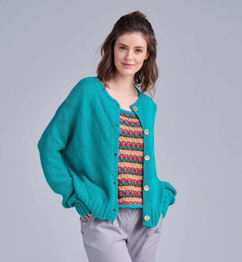 Winkende Frau in blauer Jacke und buntem Top vor neutralem Hintergrund.