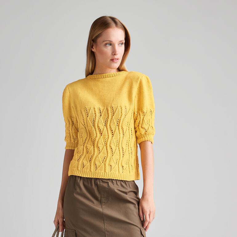 Frau in gelbem Pullover mit lockeren Ärmeln steht lächelnd vor neutralem Hintergrund.