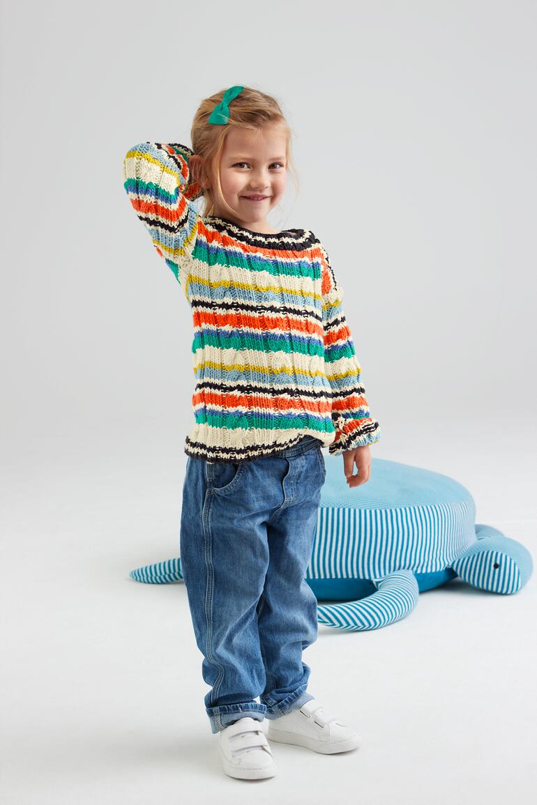 Mädchen in farbenfrohem Kinderpullover steht fröhlich und hält die Hand am Kopf.