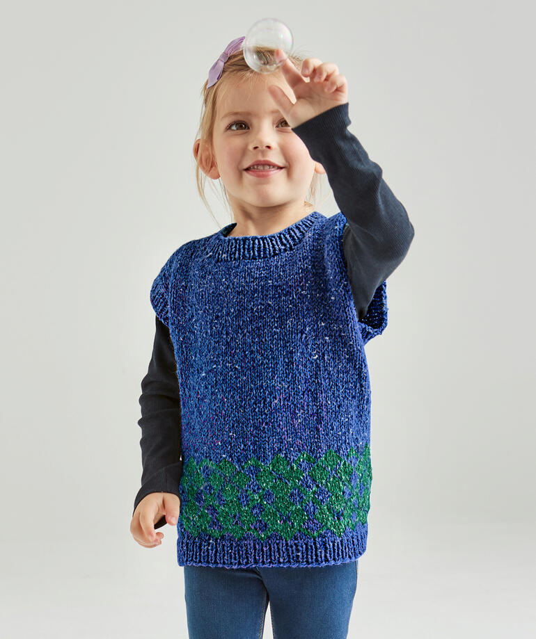 Mädchen hält eine Seifenblase in einem blauen Kinderpullunder mit grünem Muster.