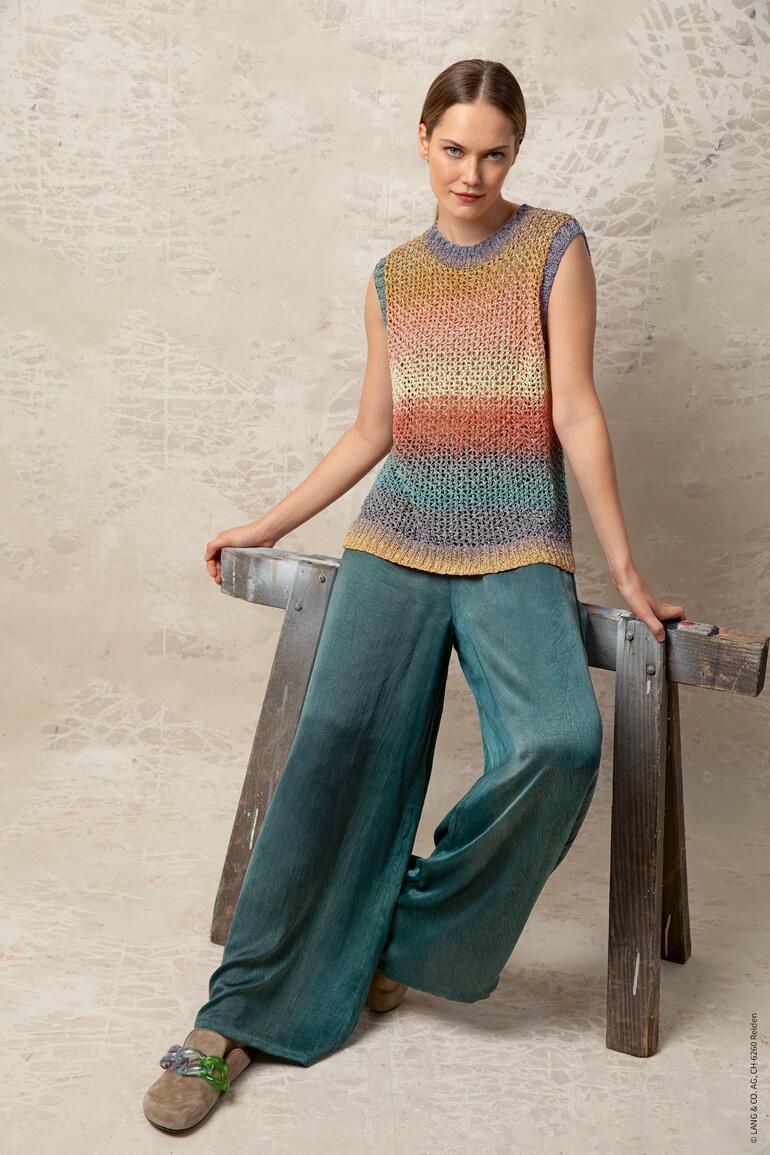 Frau in buntem Top und weiten Hosen sitzt an einer Wand auf einem Holzstuhl.