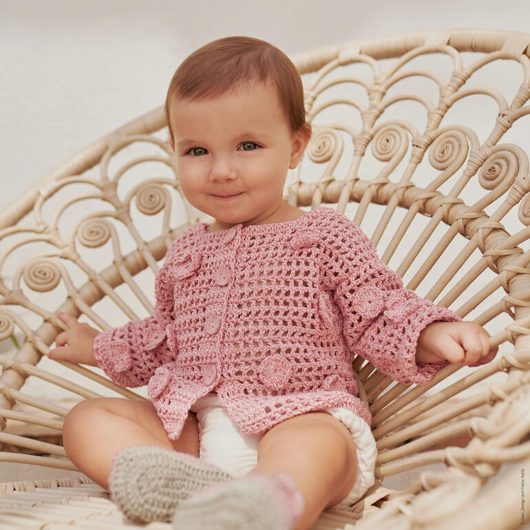 Lächelndes Baby in gehäkelter Jacke sitzt auf einem Korbsessel.