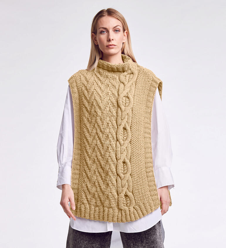 Frau in beiger Strickjacke mit Zopfmuster steht vor einem weißen Hintergrund.