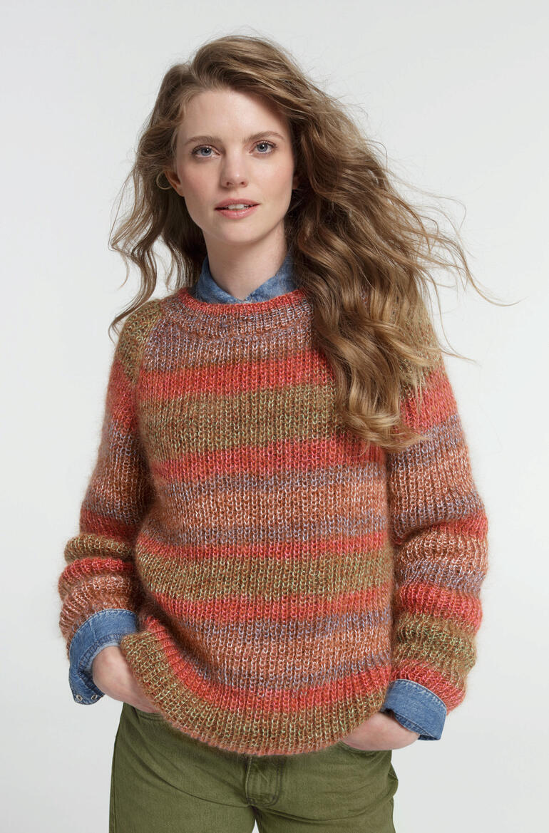 Frau in gestreiftem Pullover aus Aparta-Print steht vor hellem Hintergrund.