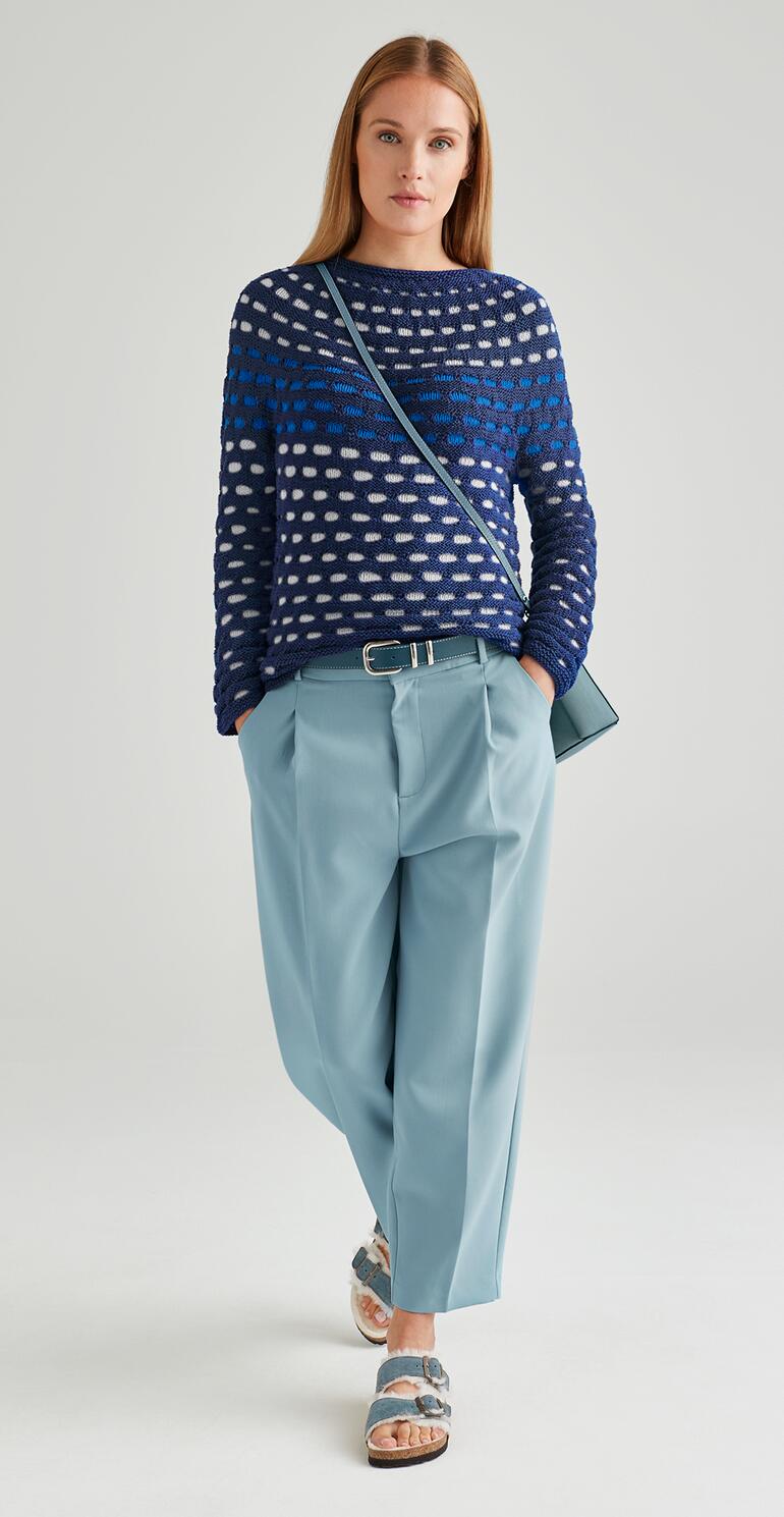 Frau in Rundpassen-Pullover mit blau-weißen Punkten steht vor neutralem Hintergrund.