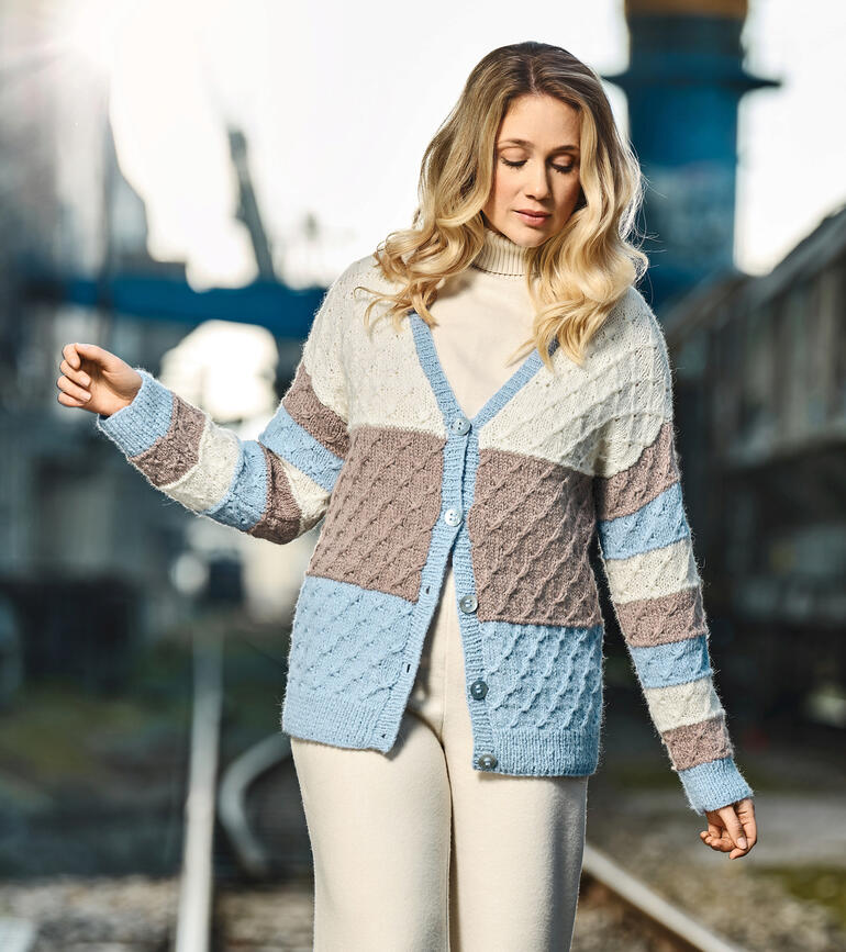 Frau in buntem, gemustertem Cardigan aus Alpaka Deluxe, steht in einer industriellen Umgebung.