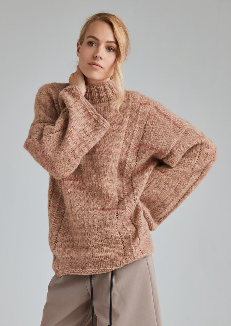 Frau trägt einen grob gestrickten Pullover in sanften Farben und steht vor einem neutralen Hintergrund.