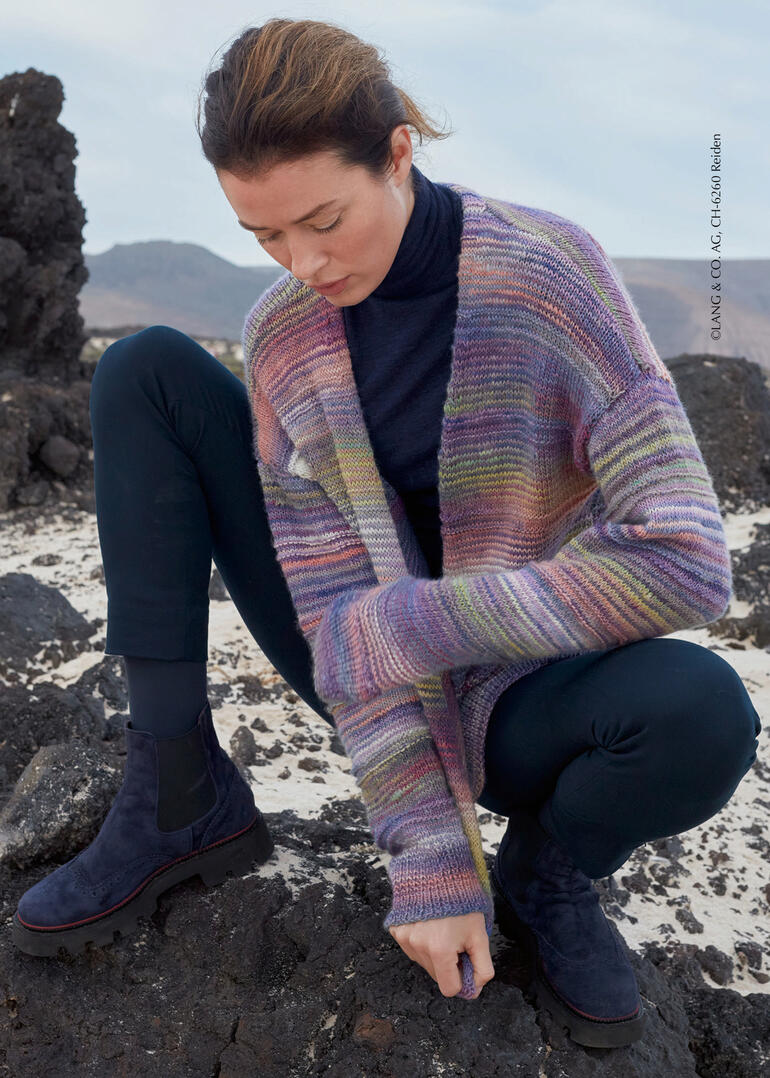 Frau in bunter Jacke aus Lovis, kniend auf steinigem Boden.