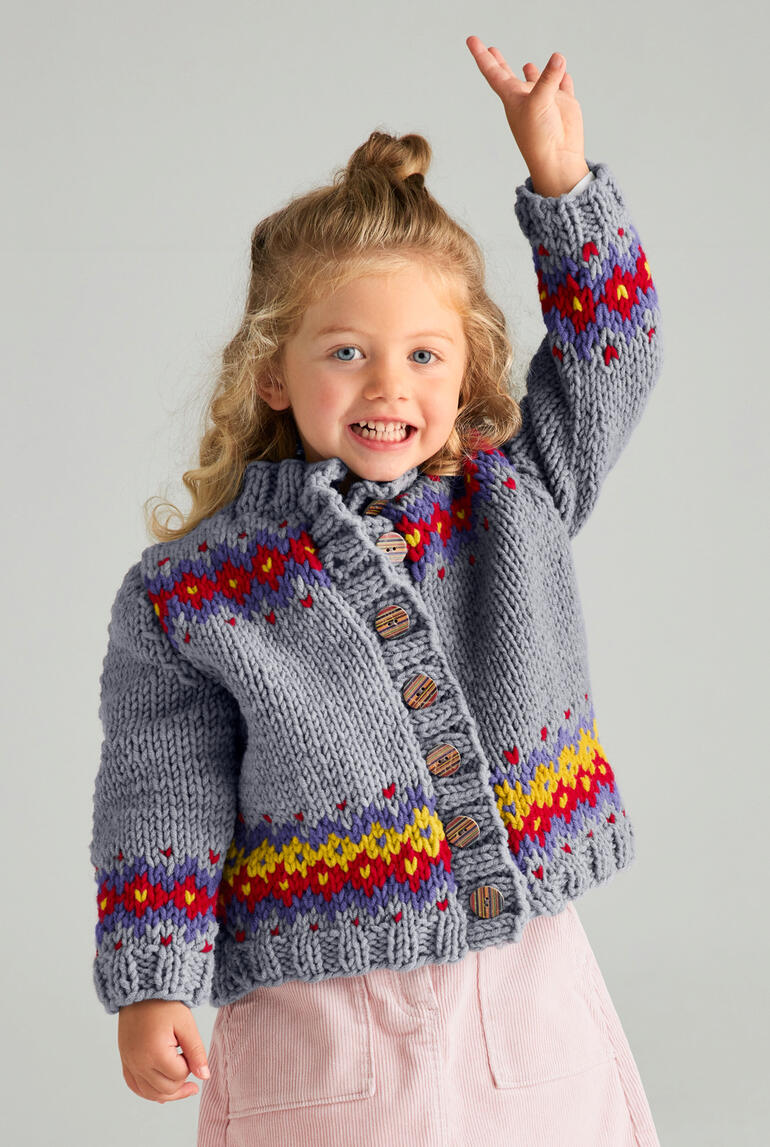 Fröhliches Kind in grauer Jacke mit buntem Muster und erhobener Hand.