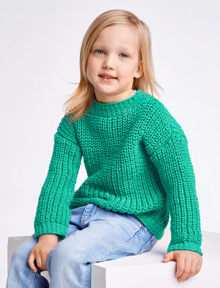 Junge mit grünem Kinderpullover aus Cotonara von Junghans-Wolle sitzt auf einem Würfel.