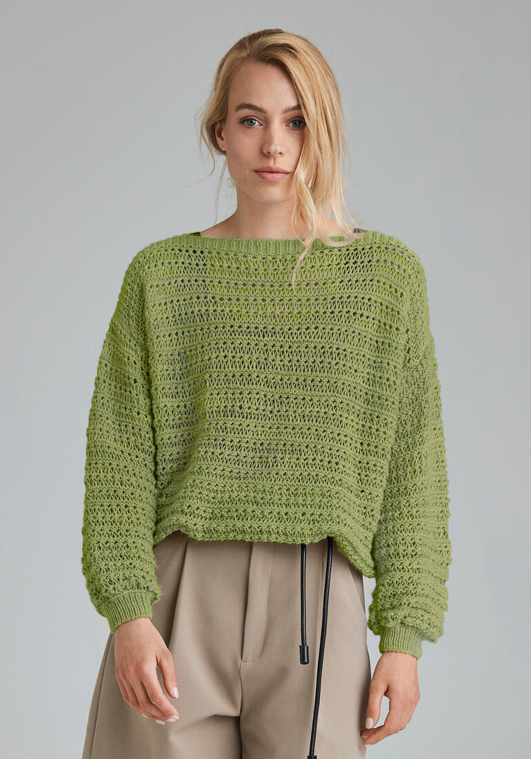 Grüner Pullover aus Babyboomer, lässig mit beigen Hosen kombiniert in einem neutralen Raum.