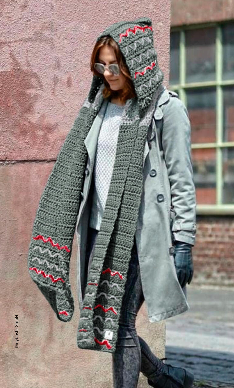 Frau trägt einen langen, gehäkelten Kapuzenschal und eine graue Jacke in urbanem Umfeld.