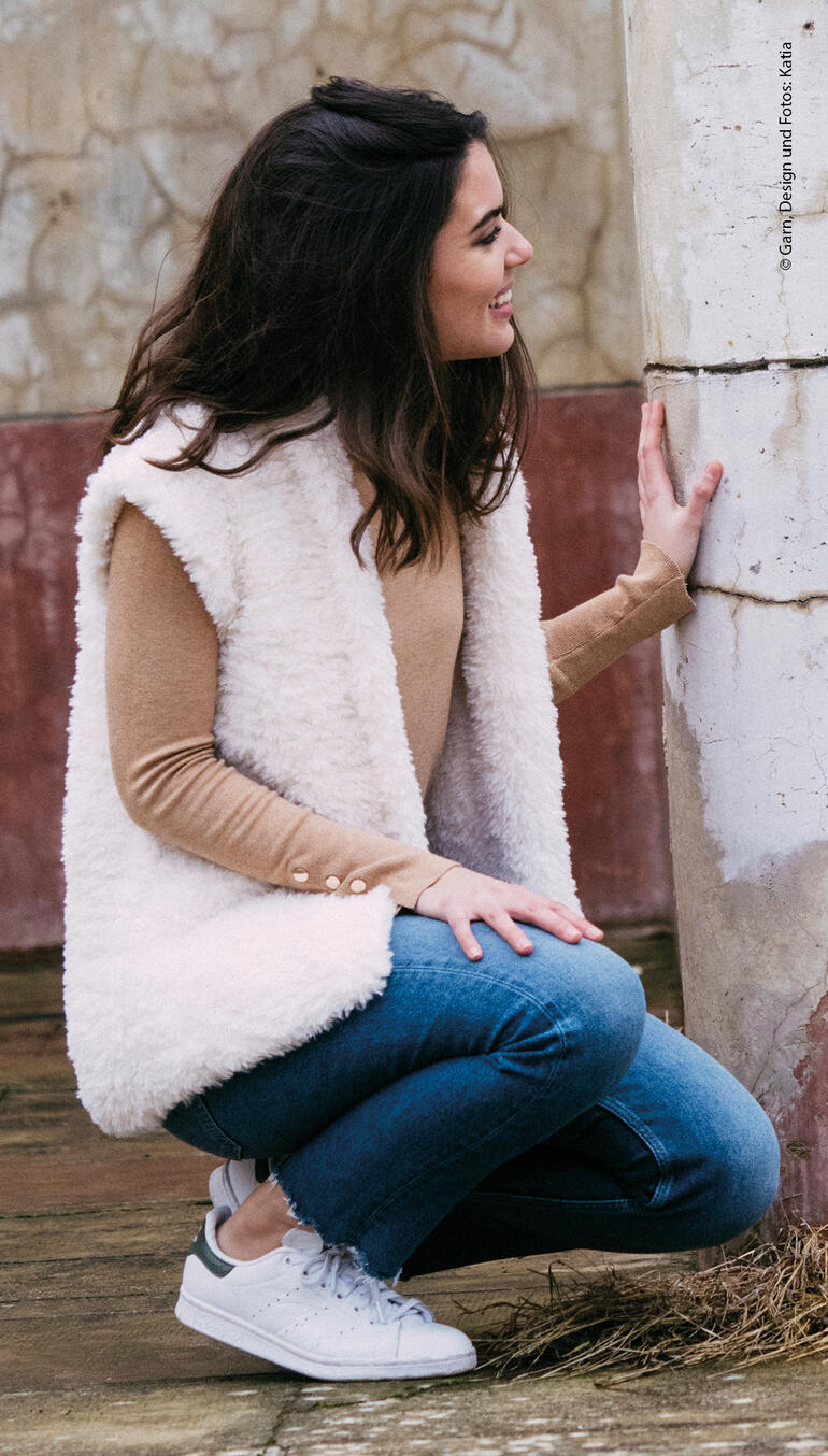 Frau kniet in einem weißen Pelzweste und Jeans, an einer Wand.