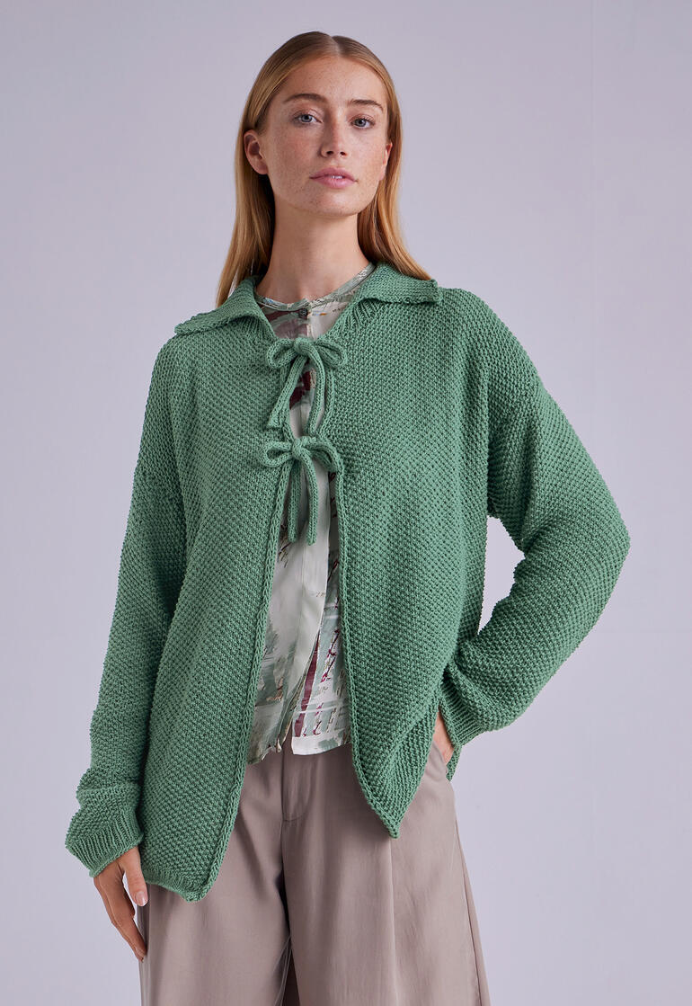 Frau in grüner Jacke mit einer Schleife, vor neutralem Hintergrund.