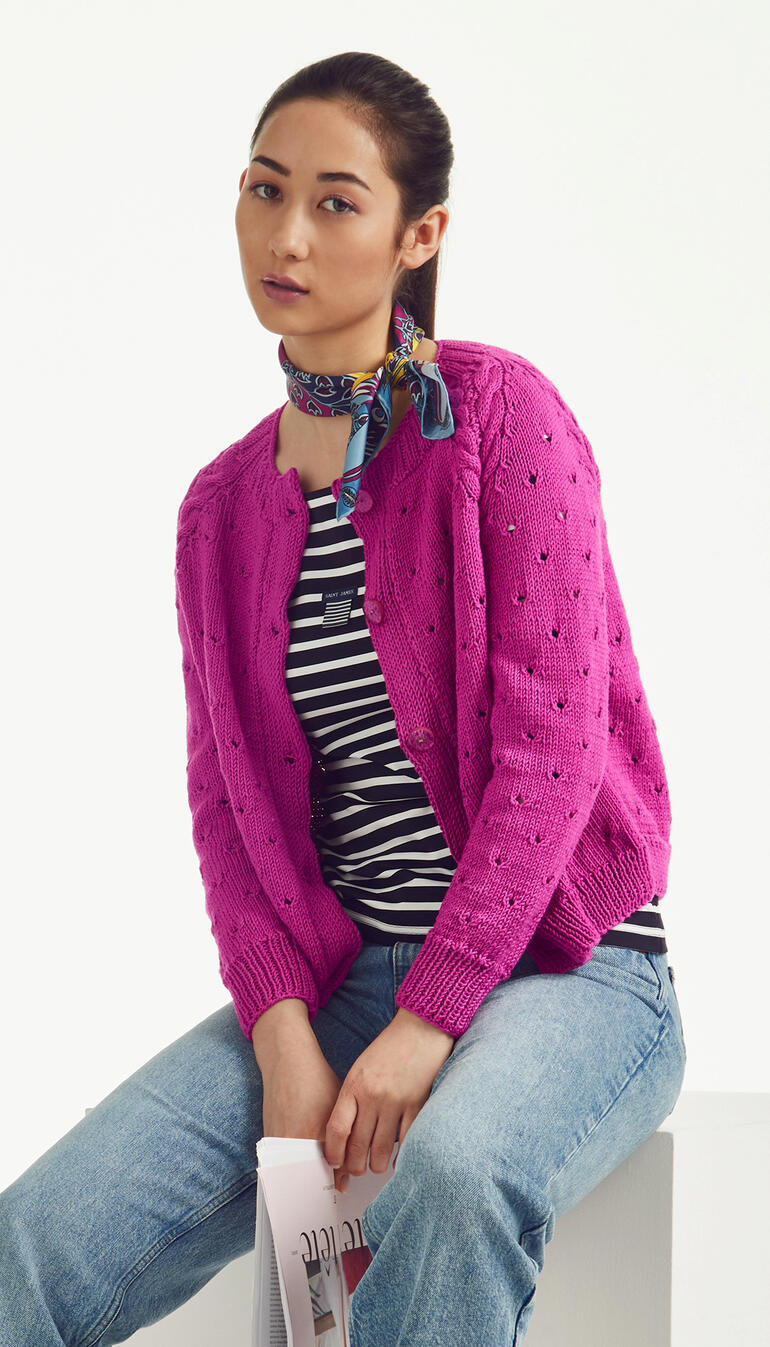 Frau in pinkfarbener Jacke mit Lochmuster sitzt auf einem Hocker.