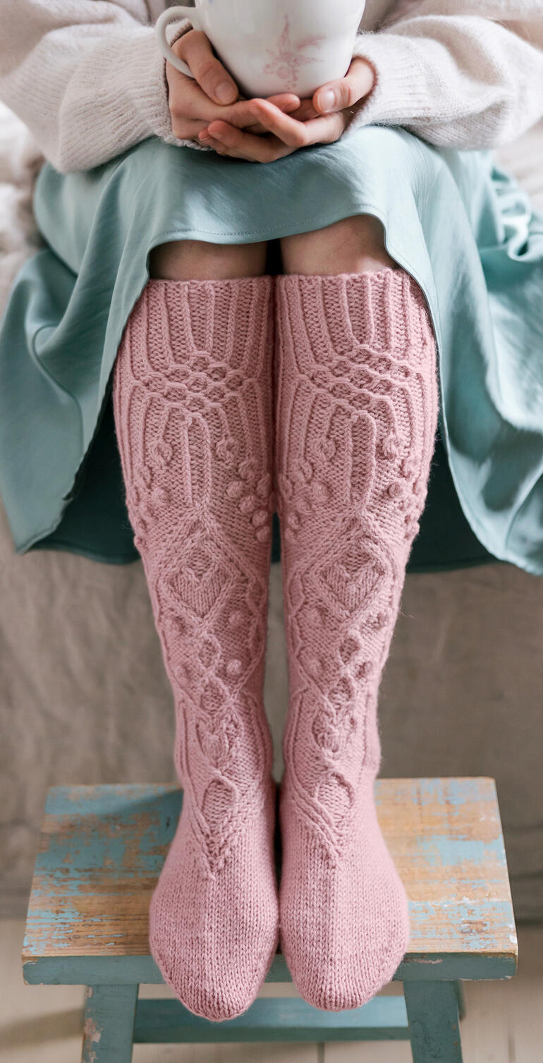 Füße mit langen, rosa gestrickten Socken auf einem Hocker.