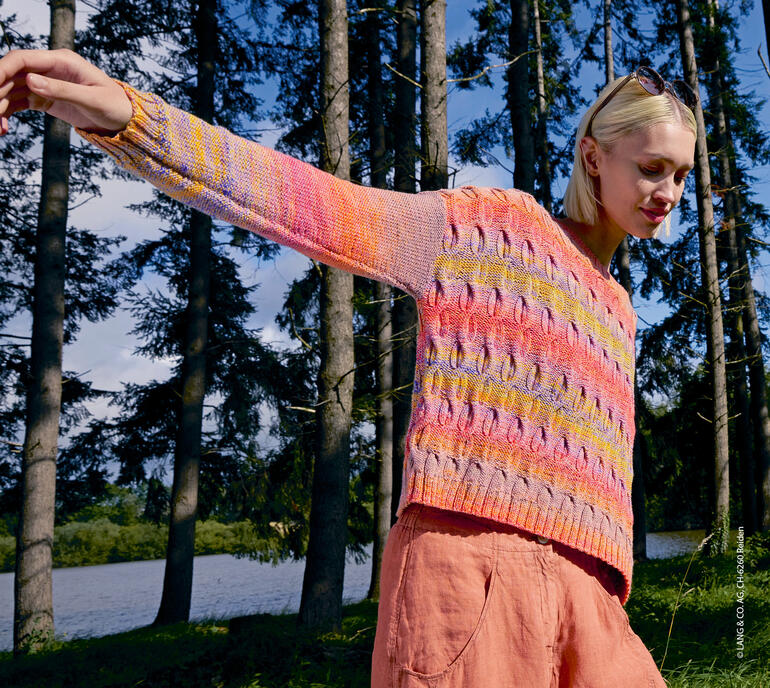 Frau in einem Pullover aus buntem Garn im Wald steht.
