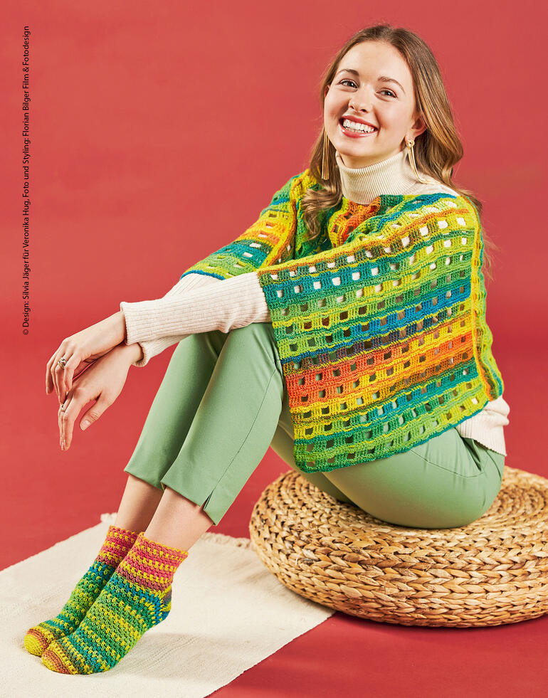 Frau in buntem Häkelponcho und passenden Socken, lächelt fröhlich auf einem Hocker.