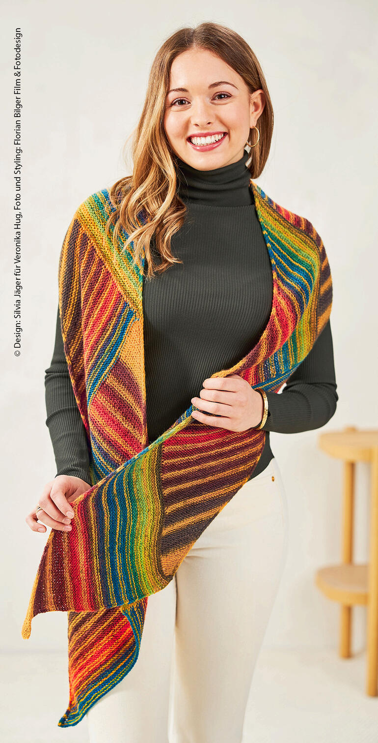 Frau mit Schal in buntem gestricktem Muster, steht lächelnd mit erhobenen Schultern.