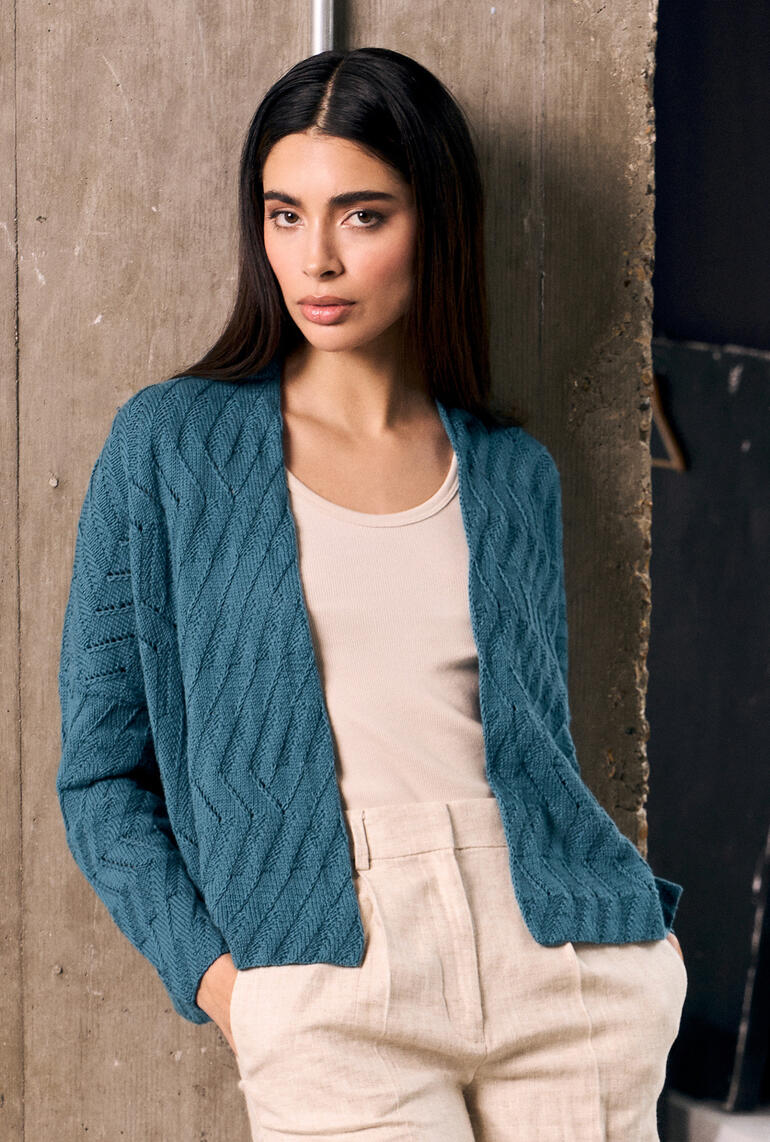 Frau in locker sitzender Jacke mit strukturiertem Muster, steht vor einer neutralen Wand.