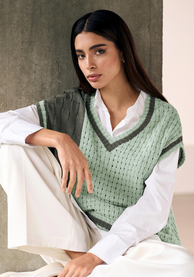 Frau in grünem Pullunder mit V-Ausschnitt, posiert lächelnd vor einer neutralen Wand.