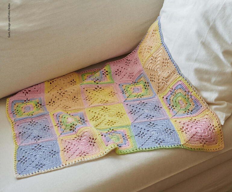 Gehaekelter Decke aus bunten Quadraten liegt auf einem Sofa.