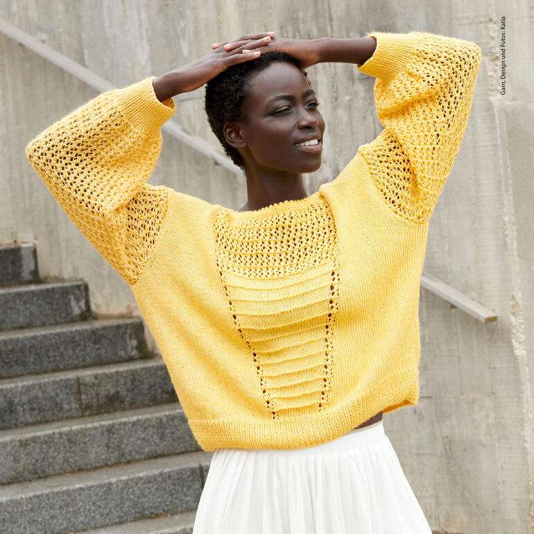 Dunkelhäutige Frau in gelbem Pullover mit Lochmuster, lächelt hinter grauer Treppe.