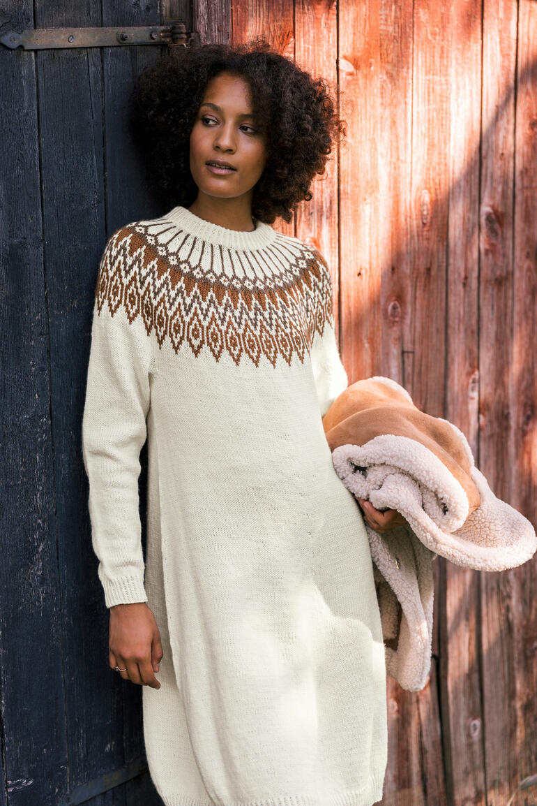 Frau in einem langen, hellen Kleid mit gestricktem Muster, hält einen Pullover in der Hand.