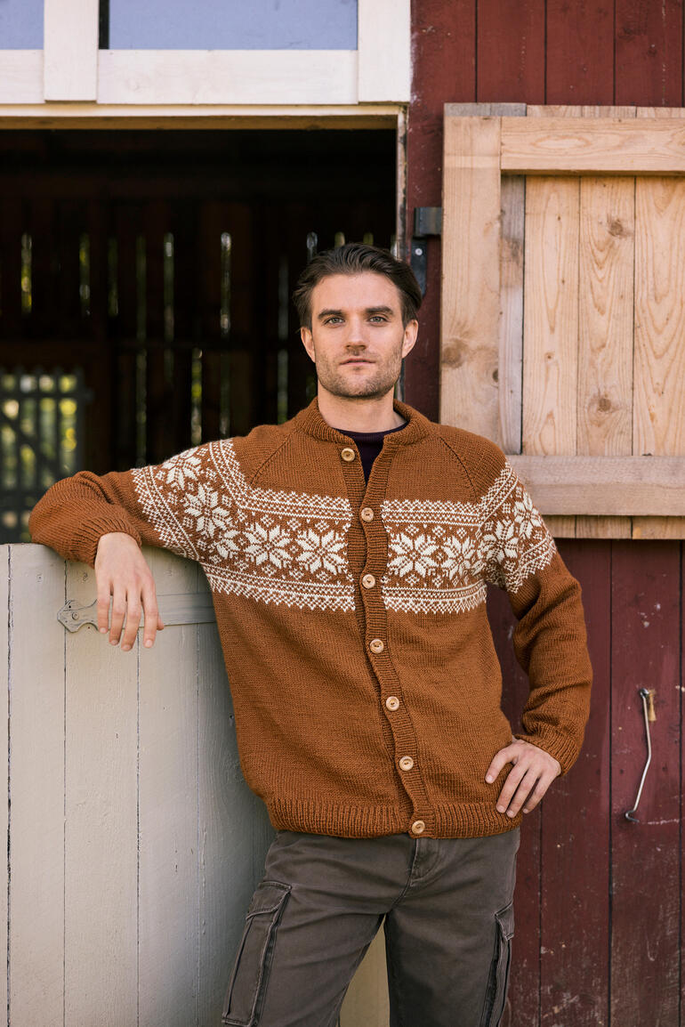 Mann lehnt an einer Wand, trägt einen braunen Cardigan mit gestricktem Muster und schaut in die Kamera.