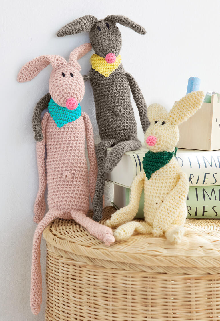 Drei gehäkelte Hasen in verschiedenen Farben, mit Halstüchern, auf einer Korboberfläche.