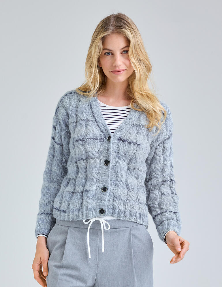 Frau in grauer, strukturierter Jacke mit Knöpfen, steht lächelnd vor einem neutralen Hintergrund.