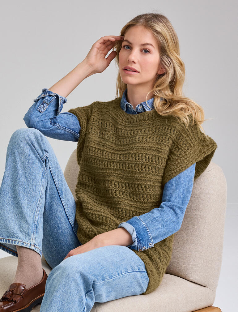 Frau sitzt auf einem Stuhl, trägt einen ärmellosen Pullunder und eine Jeans, schaut zur Seite.