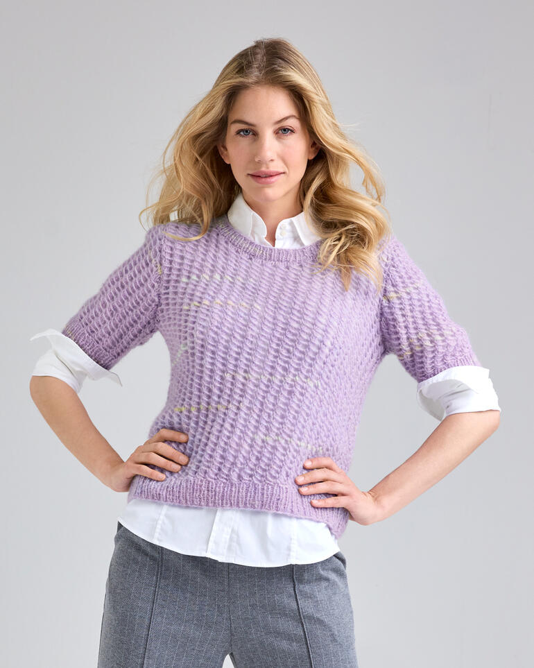 Frau in einem lila Pullover mit kurzen Ärmeln, steht selbstbewusst mit verschränkten Händen.